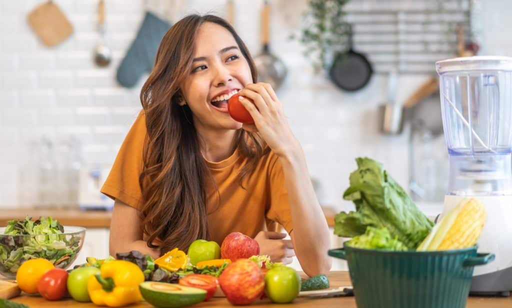 Smiling woman eating fresh vegetables in a kitchen filled with fruits and greens that support gut health.