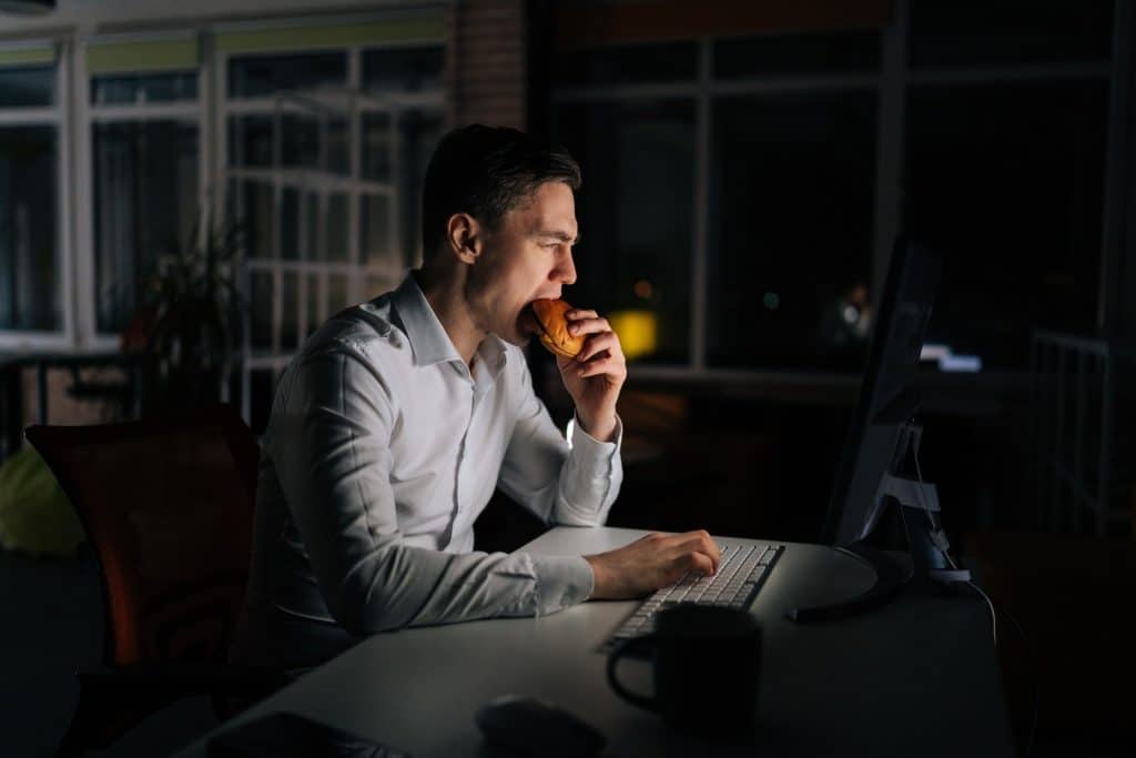 Man working late at a computer while eating fast food, showing lack of prebiotics for gut health in modern diets