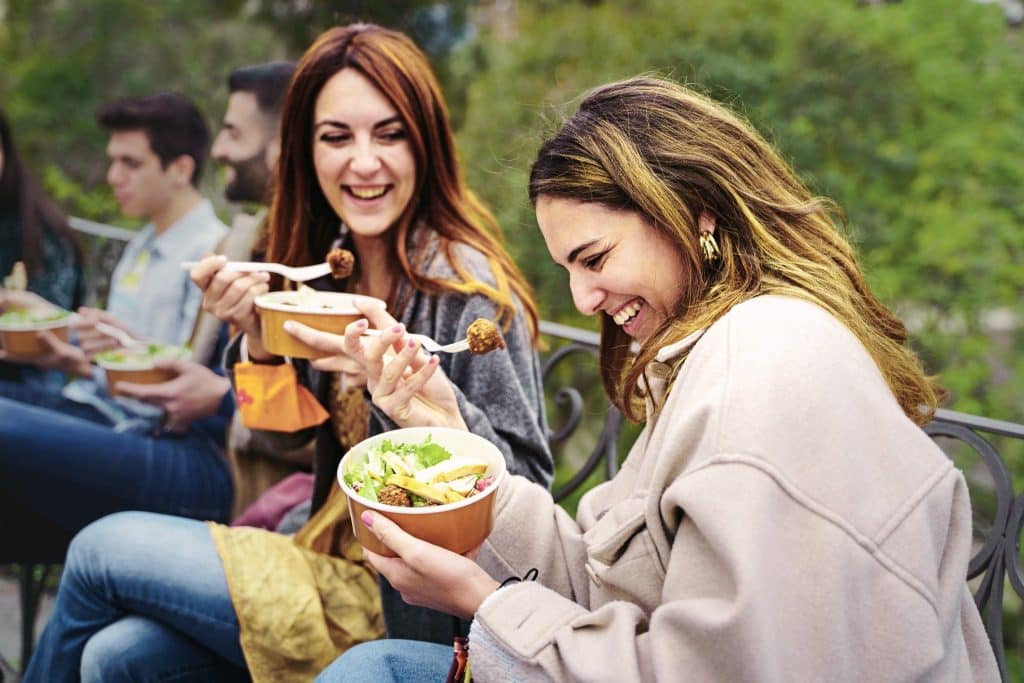 Friends sharing bowls of salad outdoors, enjoying smart snack options for busy lives in a social setting.