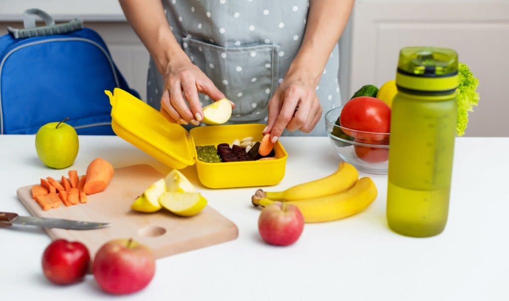 Person preparing a colorful lunchbox with apples, carrots, and bananas as part of healthy snack prep.