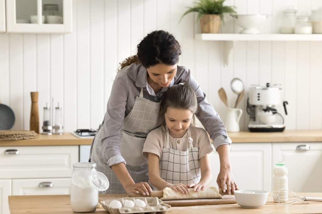 Mother teaching her daughter to roll dough in the kitchen, illustrating smart snack options for busy lives and the importance of family bonding through food.