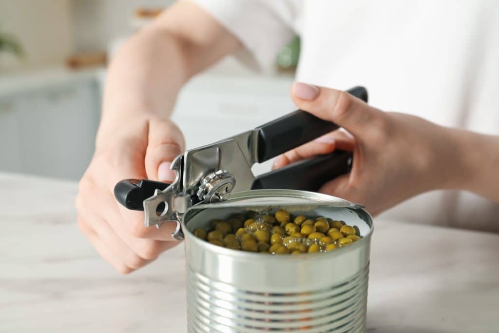 tinned fish trend at home: person opening a tin can with opener beside tomatoes, garlic, and a saucepan.